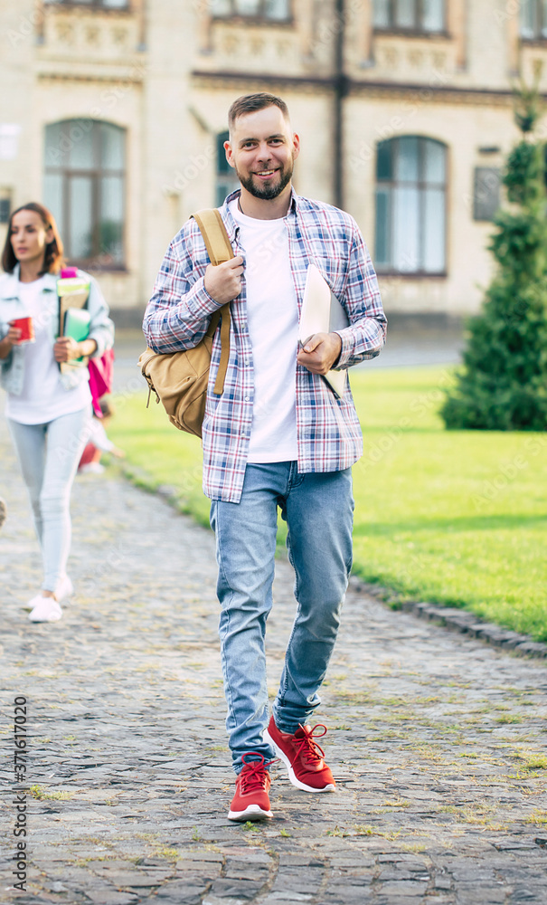 Young handsome bearded student guy in casual clothes with backpack and books in hands is walking and posing on a university buildings background Young handsome bearded student guy in casual clothes with backpack and books in hands is walking and posing on a university buildings background
