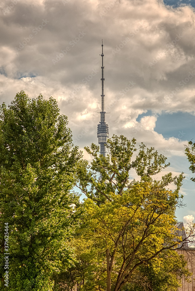 Obraz premium The main Moscow TV tower in Ostankino. Ostankino TV tower on the background of a summer storm sky through the green crowns of trees. Russia, Moscow, August 2020