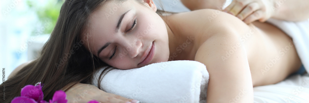 Close-up of young female resting in spa salon. Attractive brunette woman on table and massage therapist. Blooming flowers as decoration. Wellness and beauty day concept
