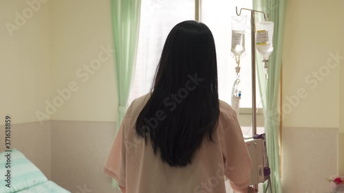 rear view woman holding drip stand is walking slowly to her bed and sitting down. camera movement lonely female patient with iv is returning to her ward