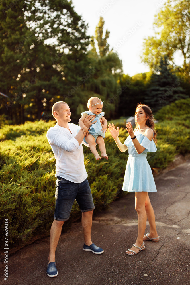 Fototapeta premium Happy mom and dad play with cute daughter, smiling, lovely parents with little baby girl at the park, caring father lift in air beautiful toddler, enjoy family moments concept