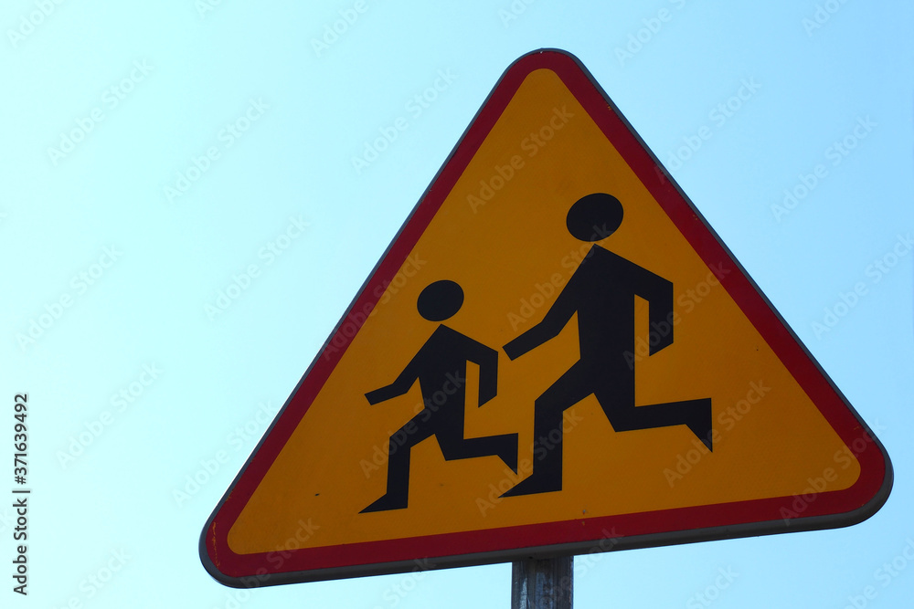 the road sign is made of metal triangular shape in orange color with the image of children against a background blue sky . Careful children the road sign is made of metal triangular shape in orange color with the image of children against a background blue sky . Careful children