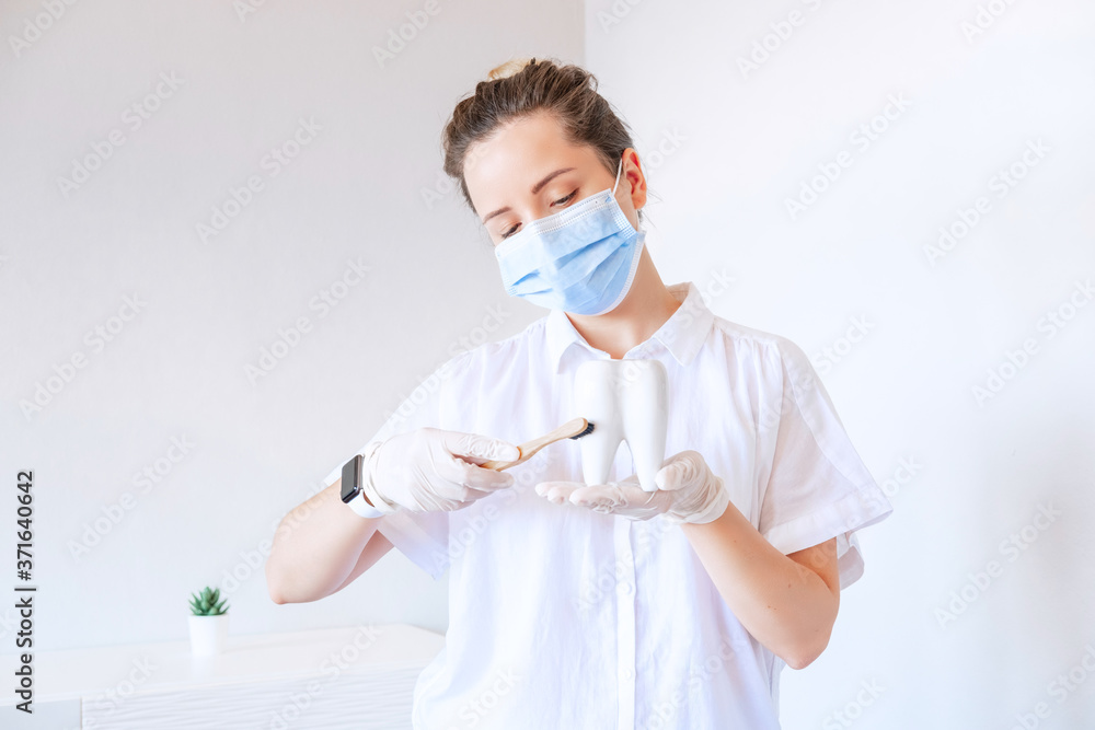 Female dentist in white uniform and protection mask, holding white tooth and bamboo toothbrush, looking at camera against stomatology interior. Medicare. Tooth cure.