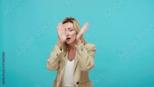 Scared terrified adult woman in business suit closing eyes and stretching hands out trying to protect herself, victim afraid of abusers. Indoor studio shot isolated on blue background