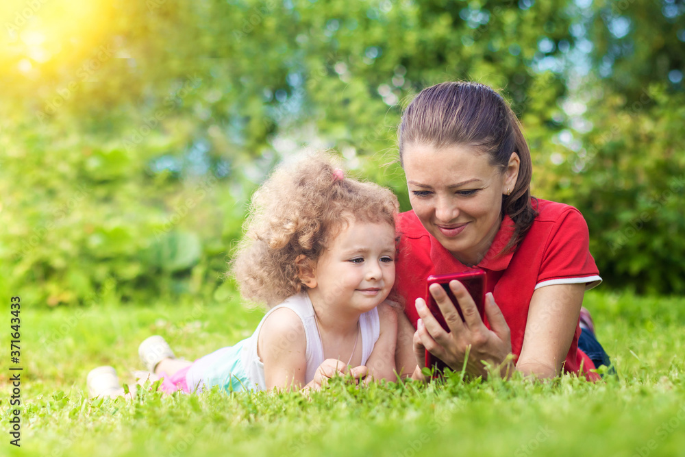 mother and daughter look at the smart phone outdoors. young woman with girl communicate online. Females smiling, showing reading on line content in gadet, lying in the grass. mother and daughter look at the smart phone outdoors. young woman with girl communicate online. Females smiling, showing reading on line content in gadet, lying in the grass.