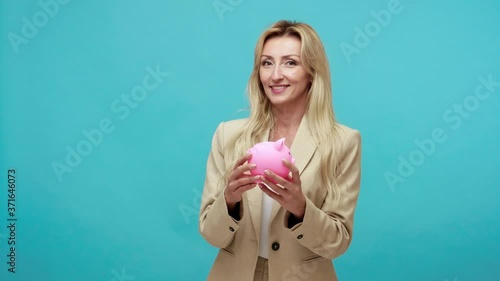 Happy smiling blond woman in business jacket holding piggy bank in hands showing thumbs up gesture, satisfied with bank deposit, crediting. Indoor studio shot isolated on blue background photo