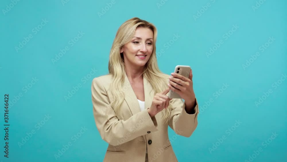 Adult business lady with blond hair using smartphone, scrolling web pages and chatting in social network, zooming with surprised face. Indoor studio shot isolated on blue background Adult business lady with blond hair using smartphone, scrolling web pages and chatting in social network, zooming with surprised face. Indoor studio shot isolated on blue background
