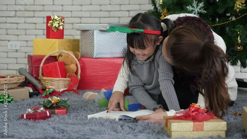 Handheld, Low angle, medium shot of Young Asian mother in sweather and little daughter with pigtails in pajamas read book on the floor, with background of christmas tree anle pile of presents photo