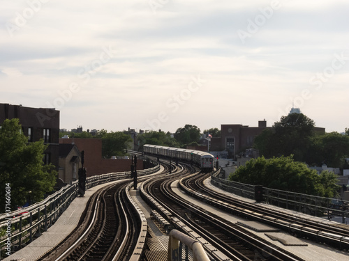 A wide-angle photo of an MTA subway train navigating the elevated tracks at 61st Street–Woodside station in Queens, New York City. Captured during daylight with a soft sky.