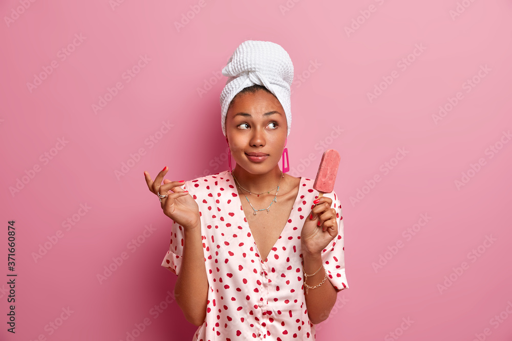 Hesitant thoughtful dark skinned woman with towel on head, enjoys eating tasty cold strawberry ice cream, has dreamy expression, wears pajama, poses against pink background. Domestic lifestyle Hesitant thoughtful dark skinned woman with towel on head, enjoys eating tasty cold strawberry ice cream, has dreamy expression, wears pajama, poses against pink background. Domestic lifestyle