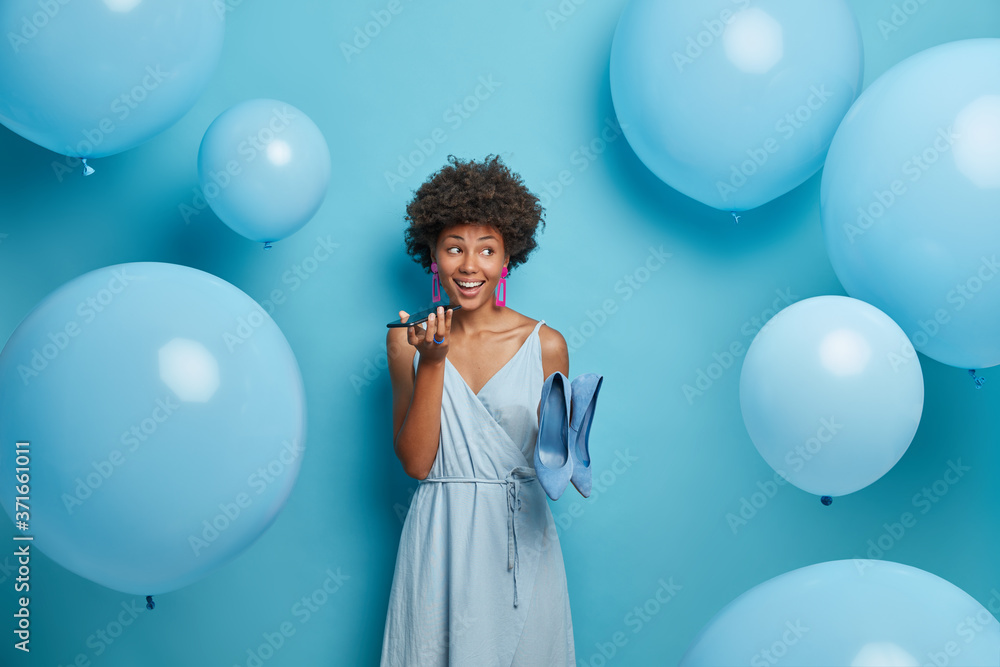 Fototapeta premium Stylish African American woman holds mobile phone near mouth, makes voice call, poses in stylish dress with high heel shoes, dresses for holiday event, stands indoor near inflated helium balloons.