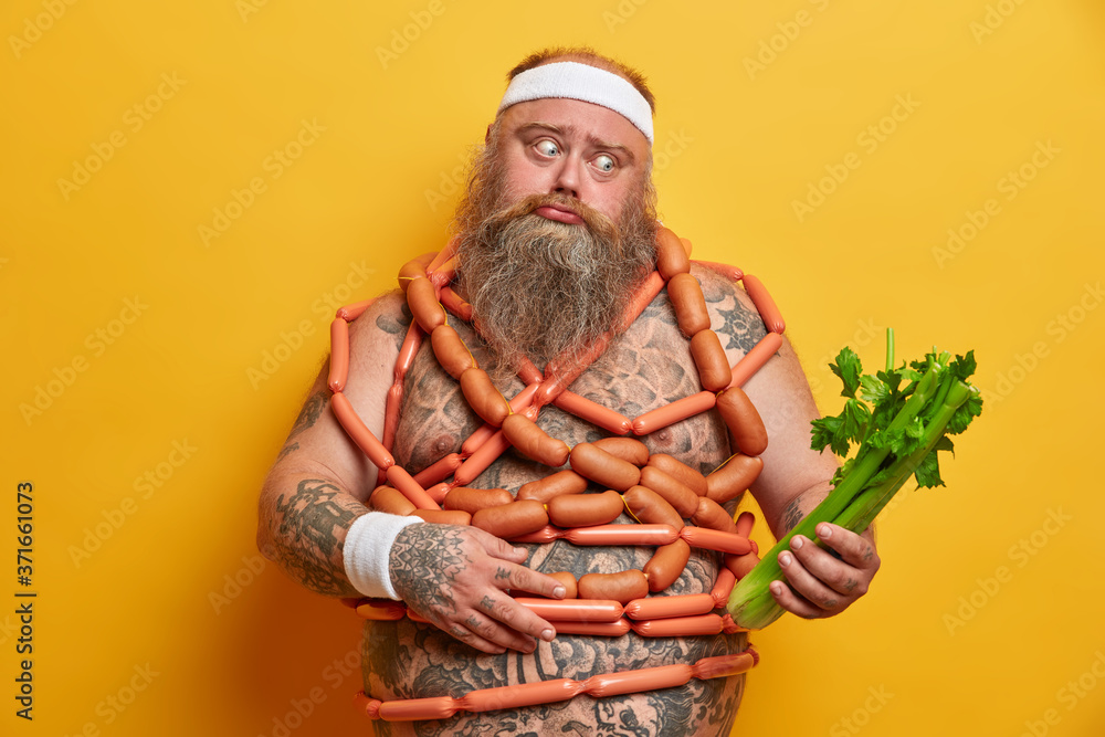 Puzzled bearded stout man chooses celery instead of sausages, controls what he eats, has healthy proper nutrition to lose weight, thick belly, tattooed body, stands against yellow background