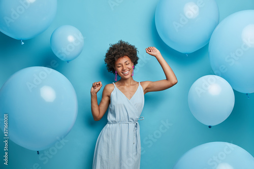 Positive smiling dark skinned woman dances carefree, keeps arms raised, wears blue fashionable dress, closes eyes, spends free time on disco party, moves against blue background with balloons