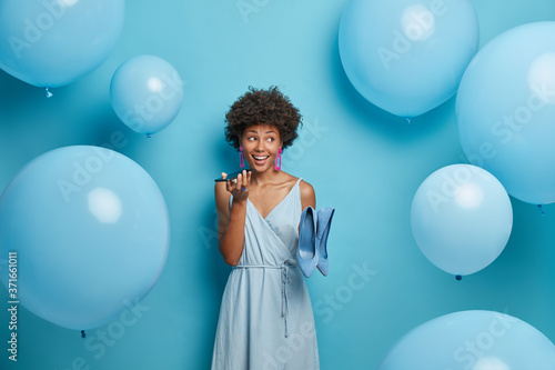 Stylish African American woman holds mobile phone near mouth, makes voice call, poses in stylish dress with high heel shoes, dresses for holiday event, stands indoor near inflated helium balloons.
