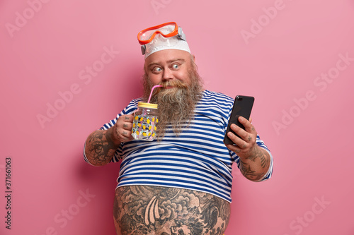 Positive fatso guy drinks fresh water, takes photo via cellphone, wears swimming snorkeling mask, undersized sailor shirt, big fat belly, poses against pink background. Summer time, technology, rest photo