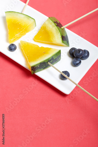 Pieces of yellow watermelon on sticks. Lie on a plate. Nearby are blueberries. A plate on a coral colored surface.