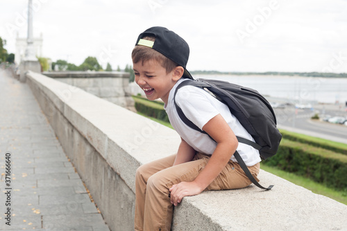 A boy of European appearance in a white t shirt and with a backpack is sitting laughing with happiness