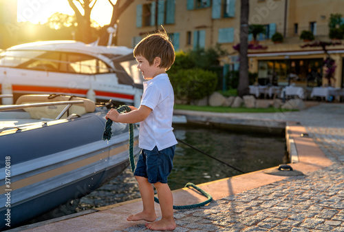 Little toddler boy mooring the boat at pier holding wet rope in hands. Child playing with big yacht at seaside. Childhood. Summer leisure activity lifestyle. photo