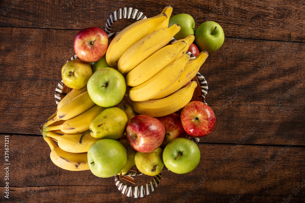fruit basket with bananas, apples and oranges on polished wood, placed on rustic wood with top view.