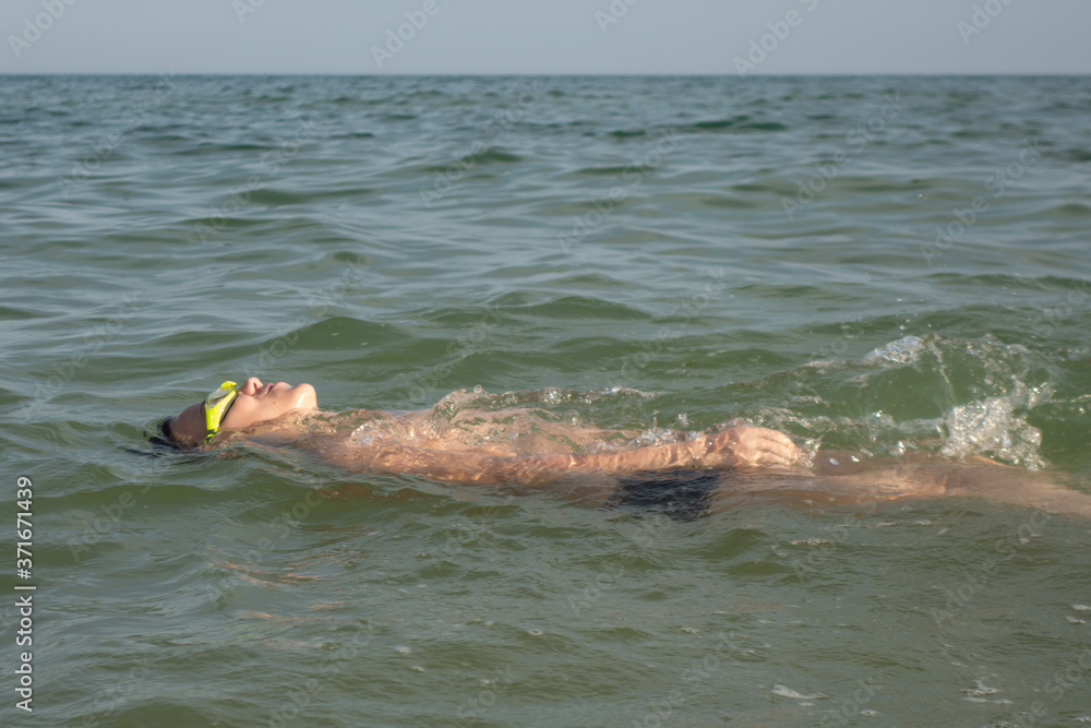 Boy 10 years old swims on his back in green swimming goggles in the sea.
