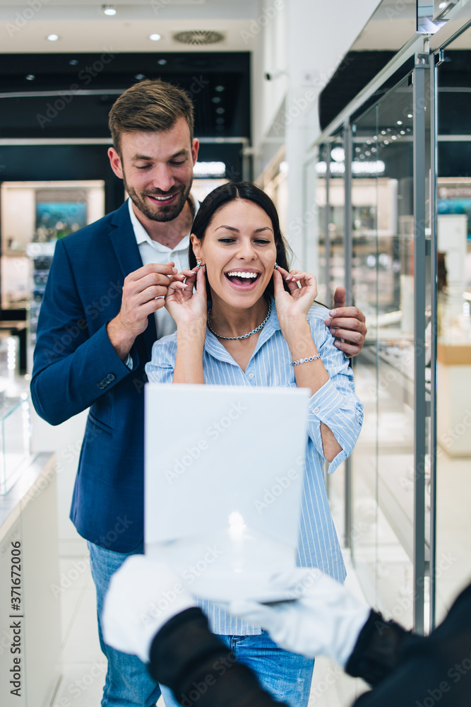 Beautiful couple enjoying in shopping at modern jewelry store. Young woman try it out gorgeous necklace and earrings.