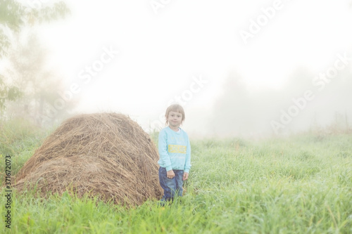 A girl stands next to a stack of dry grass against a foggy landscape