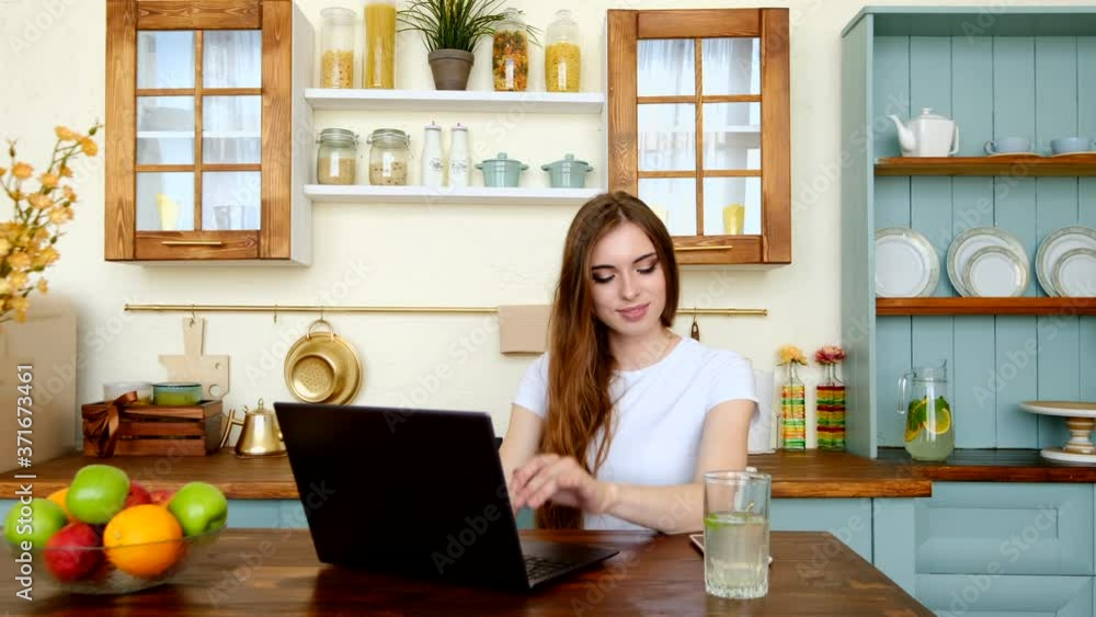 Young attractive woman with dark hair sitting at the table in the kitchen with laptop and working Young attractive woman with dark hair sitting at the table in the kitchen with laptop and working