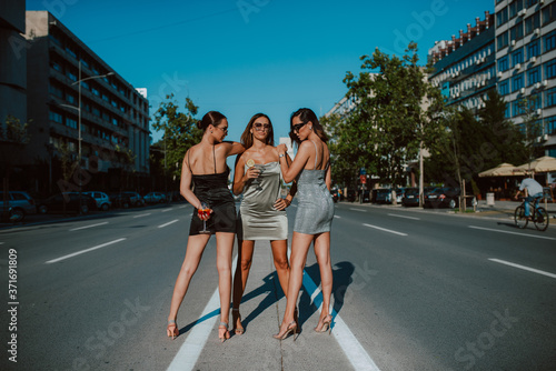 Three attractive caucasian women in summer tight dresses posing with cocktails on the road. Fashion and beauty	