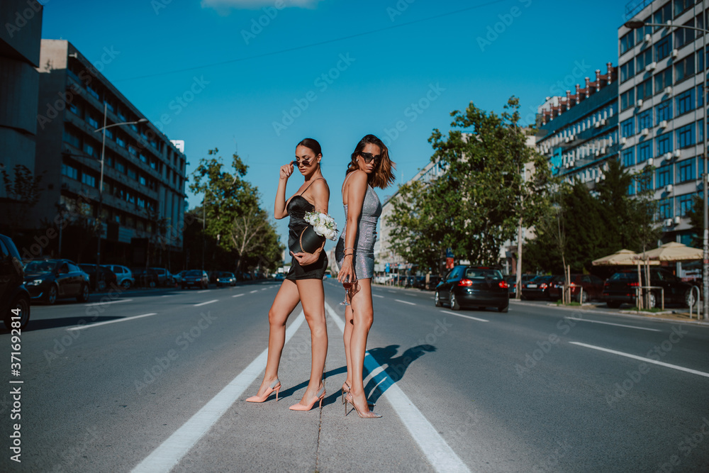 Two attractive caucasian women in summer tight dresses pose with flower boxes and cocktail on the road. Fashion and the city