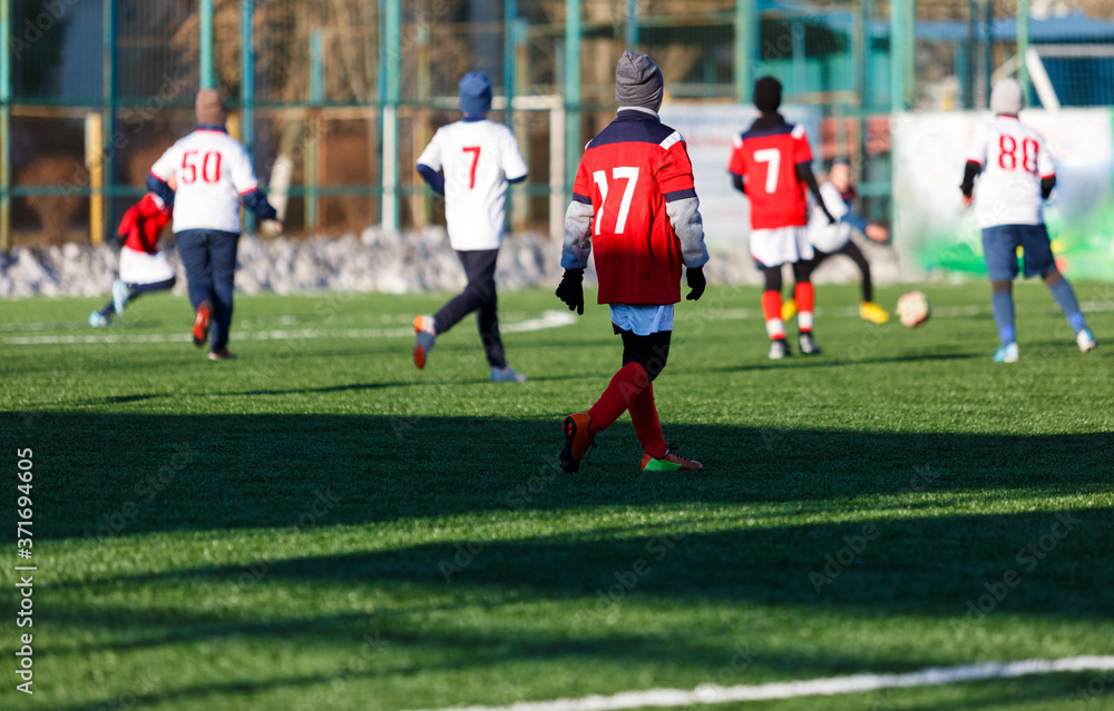 Boys in white red sportswear running on soccer field with snow on background. Young footballers dribble and kick football ball in game. Training, active lifestyle, sport, children winter activity Boys in white red sportswear running on soccer field with snow on background. Young footballers dribble and kick football ball in game. Training, active lifestyle, sport, children winter activity