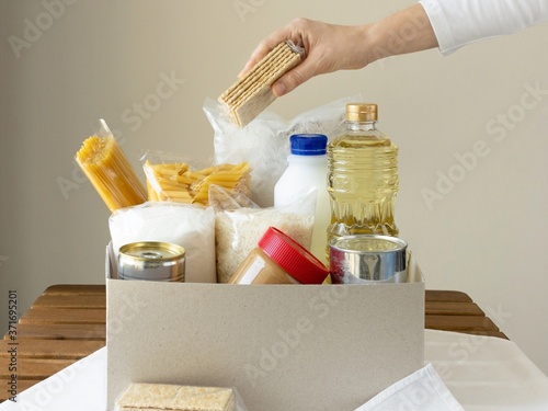 Woman's hand package food in to the box. Helping and charity concept. Oil, canned food, peanut butter, pasta, milk, sugar, rice.