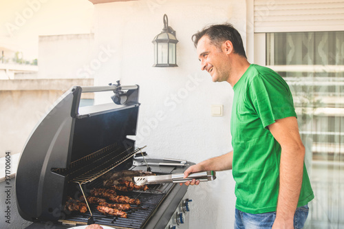 Happy Male Cooking Meat on Sticks on a Professional Grill at the Balcony