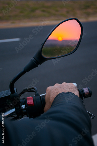 Front of a black motorcycle where you can see the handlebar with the sunset reflected in the mirror. © Mireia