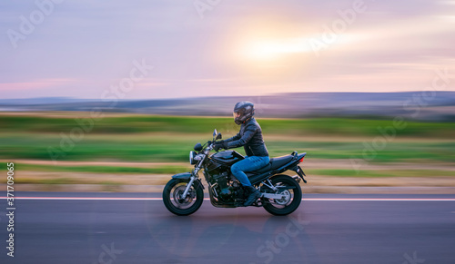 Man sitting on a black motorcycle, dressed in jeans and black jacket, moving on the road with the background out of focus.