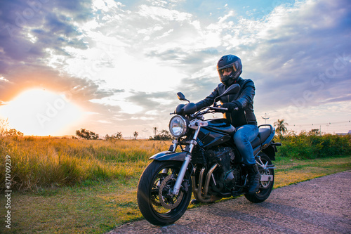 Fototapeta Naklejka Na Ścianę i Meble -  Man sitting on a black motorcycle, wearing jeans, black jacket and black helmet with a background of the sky with clouds.