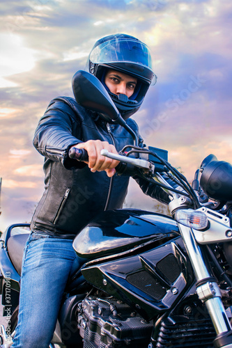 Man sitting on a black motorcycle, wearing jeans, black jacket and black helmet with a background of the sky with clouds. © Mireia