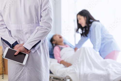 The doctor with tablet on hand for diagnosing the illness for a little girl on the hospital bed with mom beside.