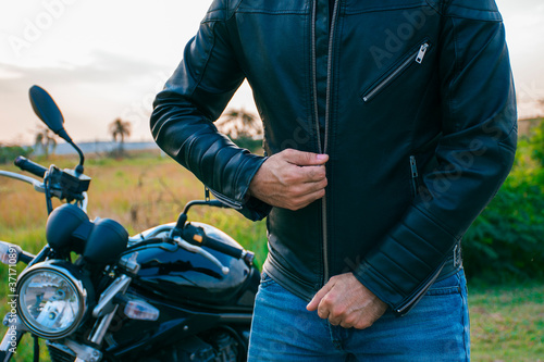 Tablou pe pânză Man whose face is not visible, dressed in jeans and a black jacket, zipping his jacket with a motorcycle behind him on the street
