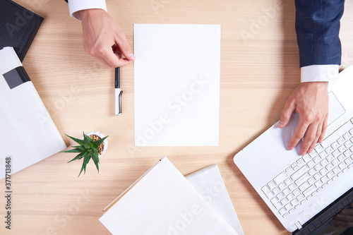 A wooden office desk with a laptop and a white sheet for recording. View from above at the hands of a businessman with a pen, ready to write. Flatley with business accessories.