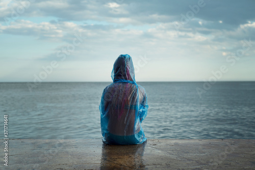Girl in pvc transparent raincoat sitting in the lonely beach, rainy day at the beach