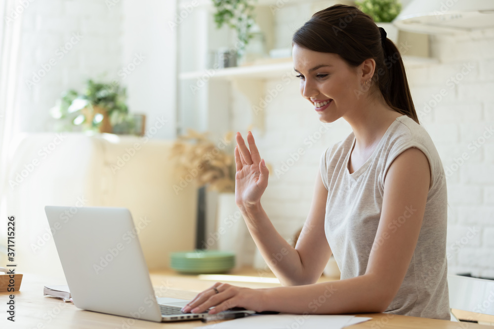 Hello there. Pleasant young female teacher or tutor sitting at home in cozy kitchen before laptop screen waving hand to camera. Student starting distant lesson, lecture or training course online Hello there. Pleasant young female teacher or tutor sitting at home in cozy kitchen before laptop screen waving hand to camera. Student starting distant lesson, lecture or training course online