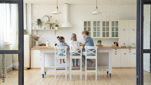 Back view of happy young mother and her little daughters sitting at kithchen table at morning having or cooking breakfast, millennial smiling nanny teaching two girls sisters to prepare food at home photo