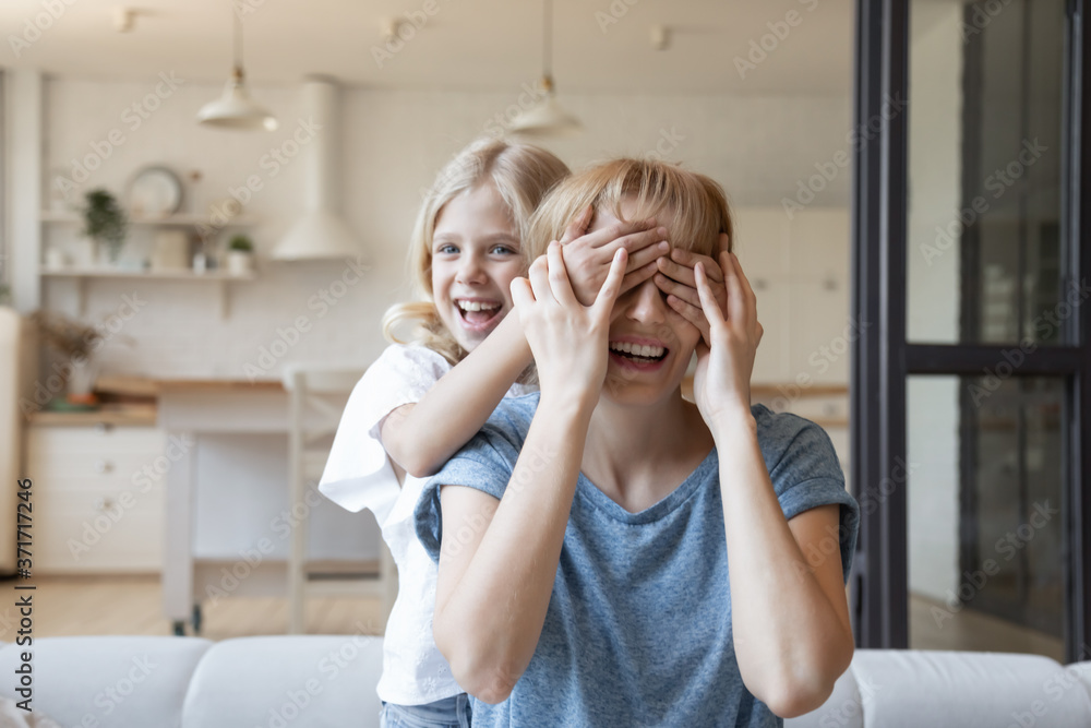 Guess who. Naughty little daughter suddenly closing eyes of her laughing mother with palms, cute funny girl playing with mommy or nanny in living room, active kid joking and making surprise for mum - obrazy, fototapety, plakaty 