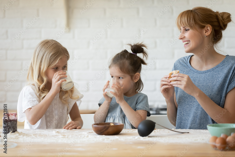 Φωτογραφία Happy family of young single mother and her two little daughters sitting at kitchen table together eating fresh homemade pastries with jam and drinking milk satisfied with result of their work