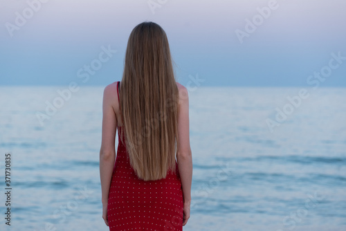 Slender girl in a red dress looks at the sea at sunset