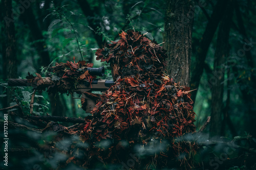 Fully covered sniper in a green forrest. Hiding behind a log. Air-soft, military, warfare.  photo