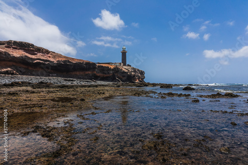 Rocky Atlantic coastline at low tide. In the background, the Punta Jandia lighthouse. Fuerteventura. Canary Islands. Spain.