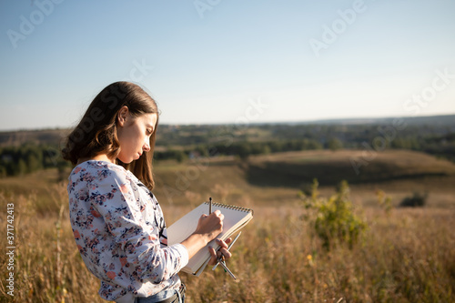 Young European woman or student is drawing in the evening outdoor in a field