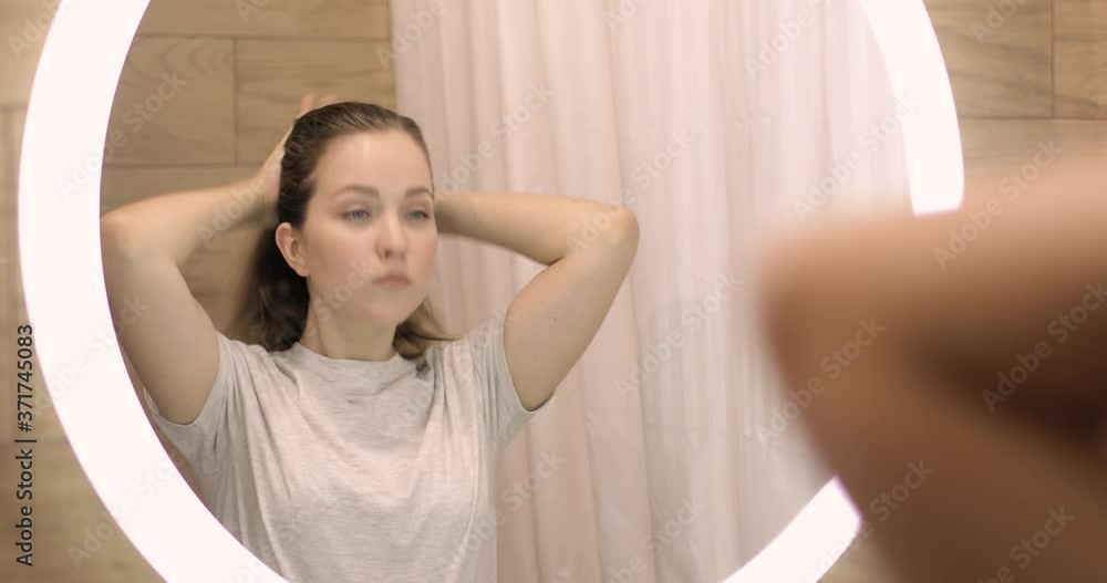 Young woman is washing applying foam cleanser on face looking at mirror in bathroom. Daily facial skincare procedure. Beauty treatment at home. She is rubbing and massaging her face. Young woman is washing applying foam cleanser on face looking at mirror in bathroom. Daily facial skincare procedure. Beauty treatment at home. She is rubbing and massaging her face.