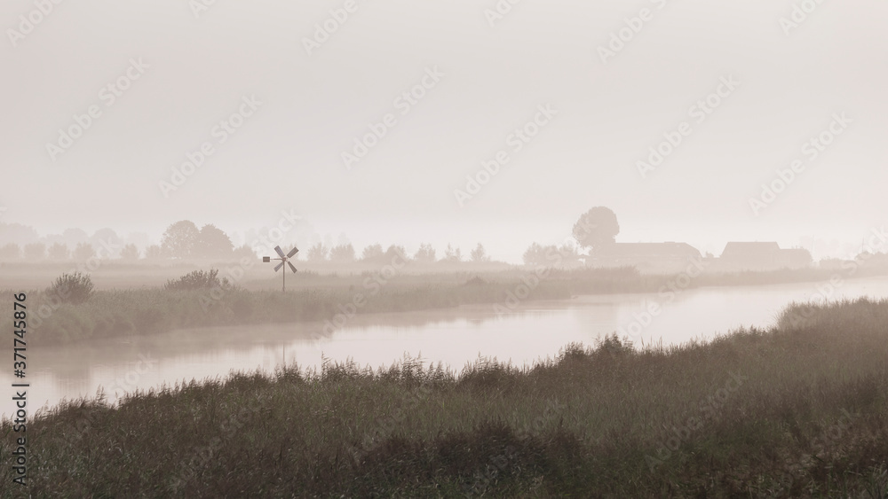 view of a dutch rural landscape with a canal and a small windmill in heavy mist, fog